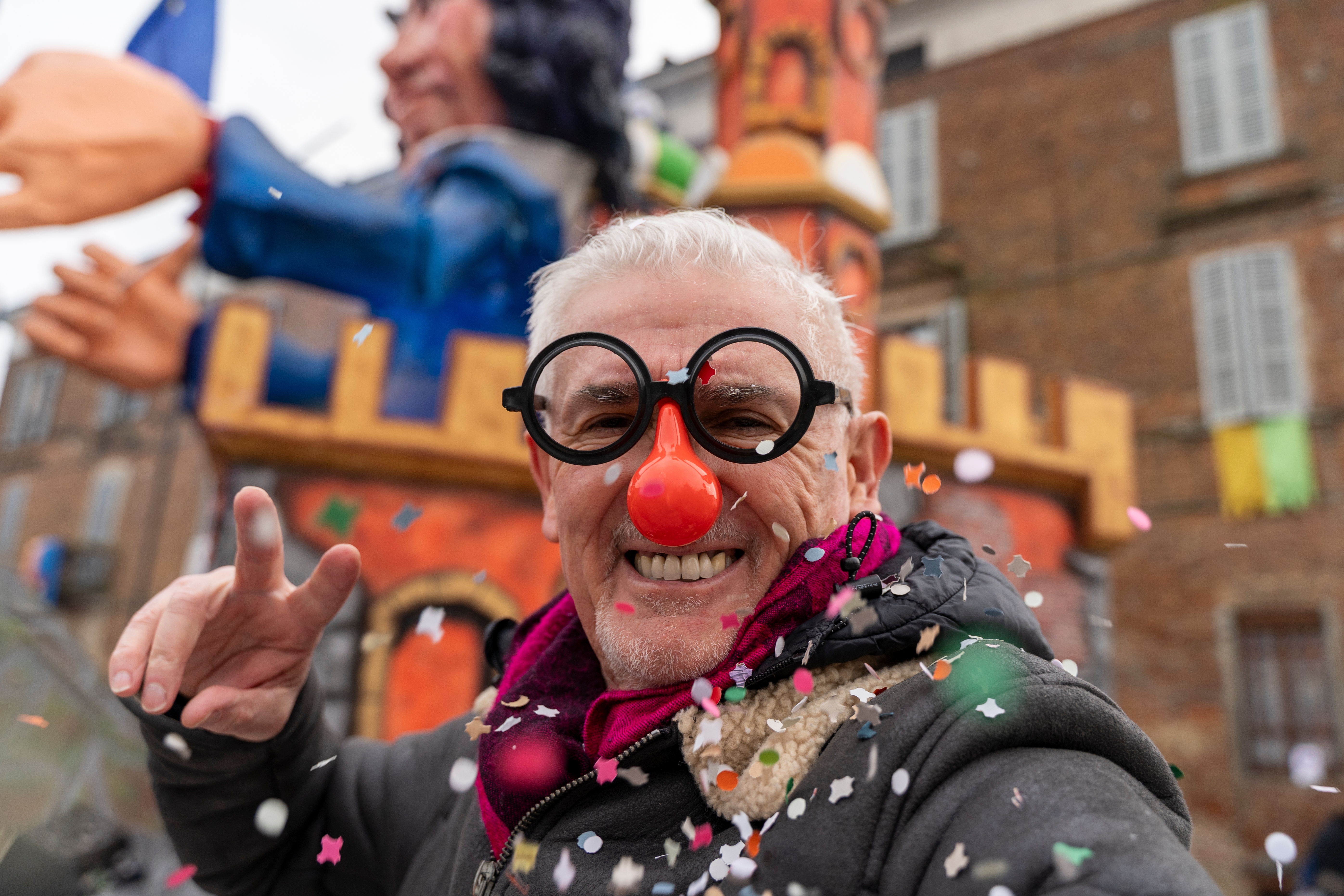 Ein lachendes Mitglied eines Karnevalsvereins mit roter Clownsnase und Scherzbrille feiert im Konfettiregen vor einem Umzugswagen.