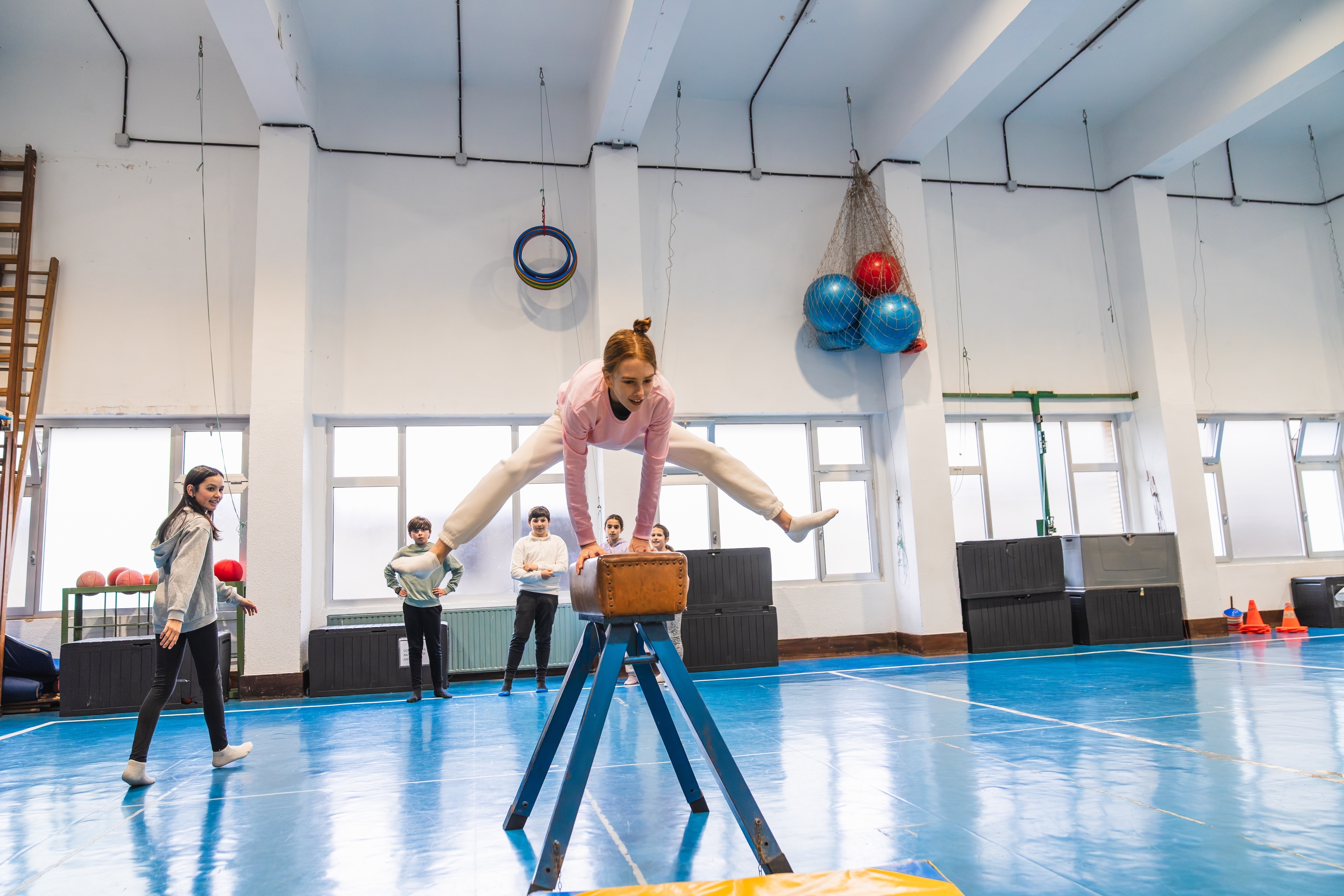 Kinder beim Geräteturnen in einem Sportverein: Ein Mädchen springt mit gegrätschten Beinen über einen Sprungbock, während andere Kinder in der Turnhalle zuschauen.